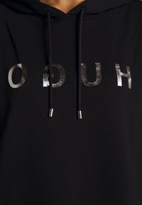 Sweat à capuche noir avec lettres argentées brillantes "HUGO" imprimées à l'envers sur la poitrine et cordons noirs avec embouts métalliques.