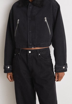 Osoba nosząca krótka czarną kurtkę z kołnierzem z baranka i srebrnymi zamkami na kieszeniach na piersi, na wysokich czarnych jeansach na jednolitym tle.