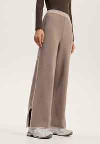 Pantaloni larghi marroni con cintura a coste, fessure laterali e texture morbida, abbinati a scarpe da ginnastica grigio chiaro.