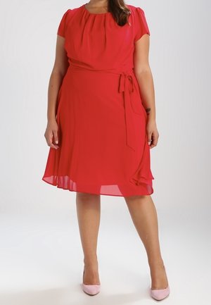 Femme vêtue d'une robe rouge longueur genou avec manches courtes et taille nouée, associée à des talons hauts rose clair, debout devant un fond uni.