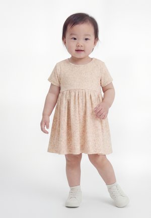 Niña pequeña de pie vistiendo un vestido rosa pálido de manga corta con un sutil patrón floral y zapatillas blancas sobre un fondo blanco liso.