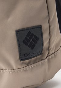 Beiger Stoff mit einem aufgenähten schwarzen Quadratsfleck, der das Columbia-Logo und den Markennamen zeigt.
