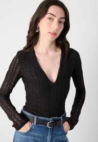 Musta pitkähihainen bodysuit syvällä V-pääntiellä, jossa on tekstuurisia geometrisia kuvioita ja lainehtivia reunoja. Yhdistetty siniseen farkkuhameeseen ja mustaan vyöhön.