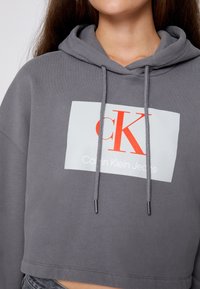 Kobieta w krótkiej szarej bluzie z kapturem z białym i czerwonym logo Calvin Klein Jeans na piersi oraz związkami w dół.
