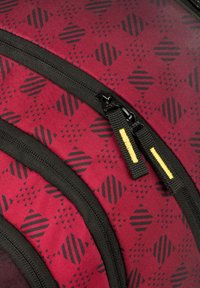 Červený látkový batoh s černým geometrickým vzorem, s vyztuženými černými zipy a žlutým akcentním stehem na táhlech zipů.