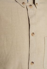 Camisa beige de botones con tejido texturizado, que presenta un cuello puntiagudo, cinco botones y un bolsillo en el pecho. Diseño clásico con costuras sutiles.