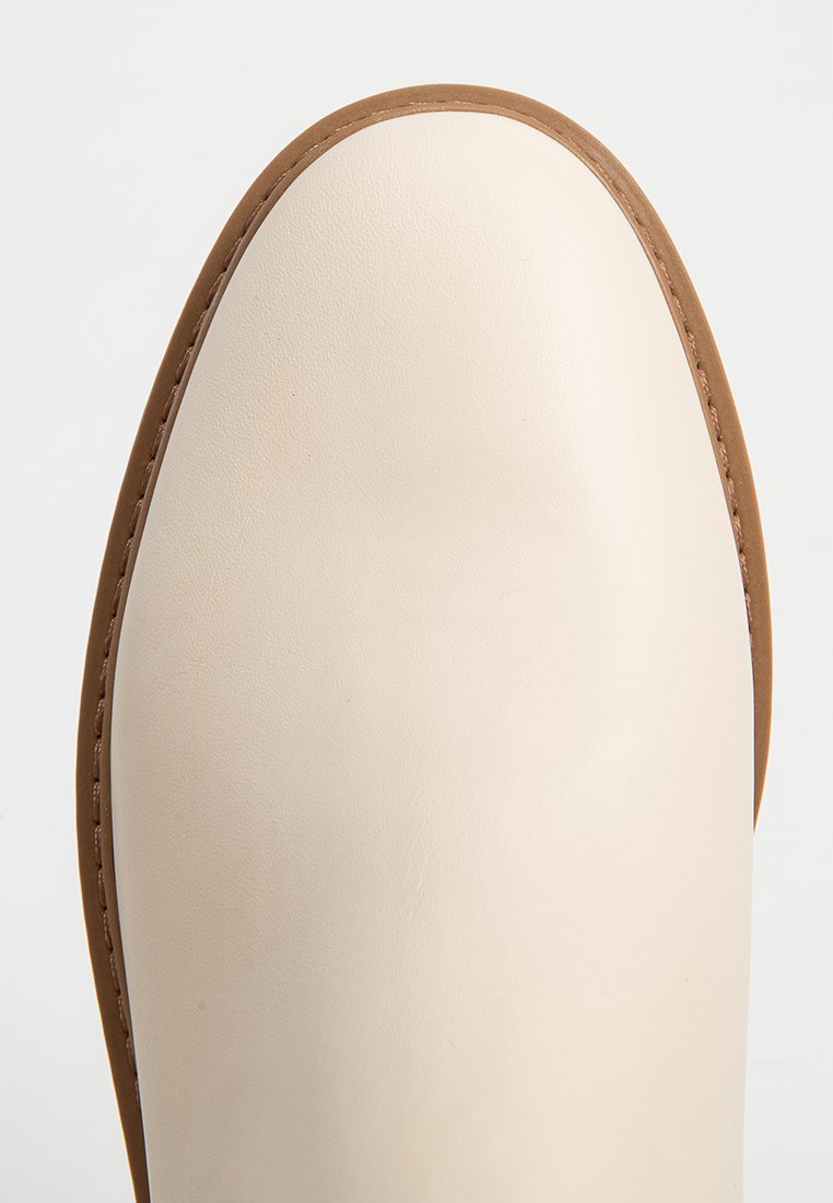Cremfarbene Lederschuhspitze mit einer glatten Textur und einem kontrastierenden braunen Gummi-Sohlenrand. Das Design ist minimalistisch mit klaren Linien.