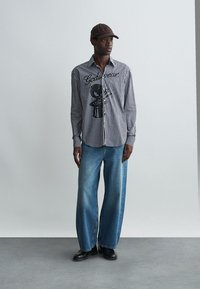 Ριγέ πουκάμισο σε μαύρο και άσπρο, με γραφική εκτύπωση, σε συνδυασμό με φαρδιά τζιν παντελόνια σε μπλε χρώμα και μαύρα παπούτσια. Φοράει καπέλο καφέ.