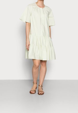 Femme portant une robe à manches courtes de couleur claire, à volants, longueur genoux avec un col rond et des sandales beiges à bride à la cheville, debout sur un sol gris.