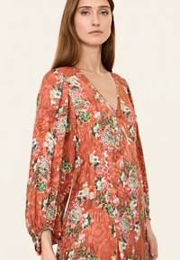 Robe à imprimé floral dans des tons rouille avec un décolleté en V et des manches bouffantes. Caractérisée par des fleurs colorées sur un tissu texturé ; ourlet arrondi et profondes poches.