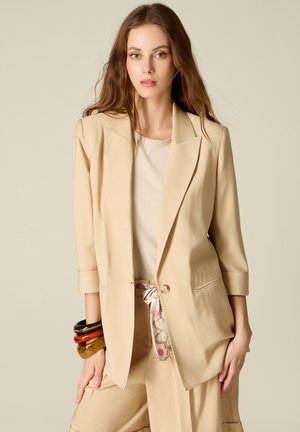 Kobieta ubrana w beżową marynarkę i spodnie z wzorzystym paskiem, stoi z jedną ręką na udzie i nosi kolorowe bransoletki.