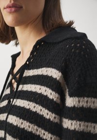 Schwarz-beige gestreifter Strickcardigan mit Kragen, strukturierter Webart, Schnürdetail am Ausschnitt und offenem Maschenmuster.