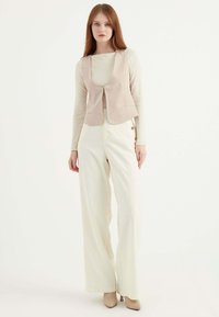 Beige-farbenes, tailliertes Vest mit V-Ausschnitt, kombiniert mit hochgeschnittenen, weißen, weit geschnittenen Hosen. Glatte Stofftextur, minimalistisches Design, Riemchen-Heels.