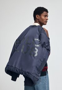 Granatowa kurtka bomberka o satynowym wykończeniu, z dużą grafiką "POLO CLUB" na plecach, ściągaczami przy rękawach i luźnym krojem.