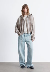 Brown plaid hooded shirt with buttons, worn over a white long-sleeve top. Light blue wide-leg jeans and black shoes complete the outfit.