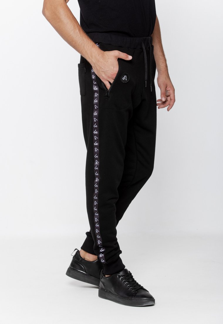 Czarne dresy z elastycznym pasem na sznurek i bocznymi kieszeniami. Białe logo oraz wzór wzdłuż zewnętrznych szwów; zwężający się krój nogawek.