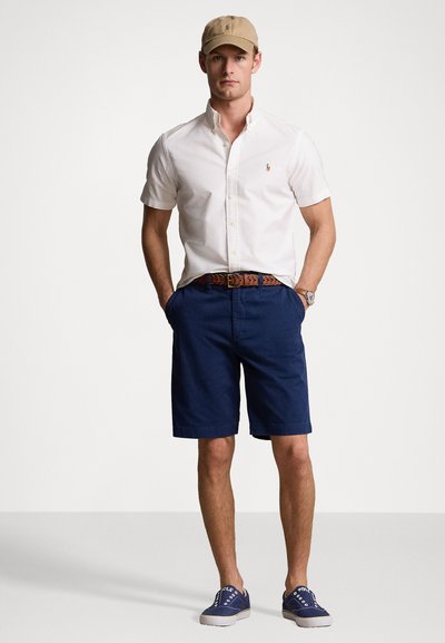 Men's outfit featuring a white short-sleeve button-up shirt, navy shorts, brown belt, and navy sneakers. Cap is light tan with logo detail.