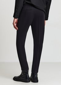 Schwarze maßgeschneiderte Hose mit schmaler Passform, die eine glatte Textur und subtile Gesäßtaschen aufweist und mit schwarzen Lederstiefeln kombiniert wird.