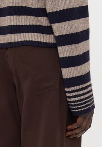 Stripet strikkegenser i marineblå og beige, med ribbet krage og mansjetter. Kombinert med brune teksturerte bukser som viser lomme detaljer.