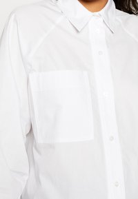 Camicia bianca a maniche lunghe con colletto classico, dotata di una tasca sul petto e una texture in cotone liscia.