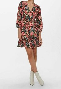 Žena stojící s překříženýma nohama, oblečená v květovaných zavinovacích šatech po kolena s 3/4 rukávy a světle zbarvených kotníkových botách.