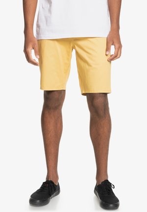 Homme portant un short jaune jusqu'aux genoux, un t-shirt blanc et des baskets noires, debout devant un fond blanc.