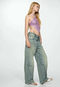 Top halter a maglia viola con un design triangolare abbinato a jeans larghi blu chiaro. La modella sta in piedi con i capelli lunghi, mostrando un ventre scoperto.
