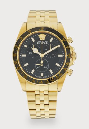 Montre en inox doré avec un cadran noir, sous-cadrans chronographe et motif clé grec. Elle est dotée d'un bracelet en métal lisse et d'une finition polie.