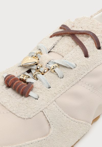 Gros plan d'une basket beige avec un tissu texturé, des lacets marron et gris clair, des perles dorées et un charme en forme de cœur doré sur les lacets.