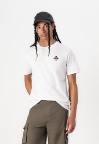 Hvit bomullst-skjorte med et lite blomsterdesign og tekst på brystet, sammen med olivengrønne cargo-shorts og en svart caps.