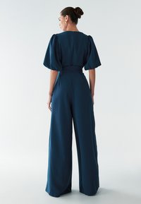 Marineblauwe jumpsuit met wijde pijpen, korte pofmouwen, een ceintuur in de taille en een rits aan de achterkant. Soepele stof met minimale texturen, geschikt voor diverse gelegenheden.