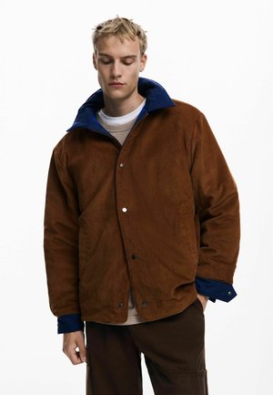 Brown corduroy jacket with a blue collar, featuring snap buttons and a relaxed fit. Worn over a light beige shirt with a round neckline.
