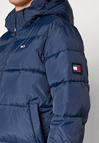 Marineblå pufferjakke med en blank finish, der har lynlås, hætte og et logo-patch på ærmet. Tekstureret, quiltet design med lommer.