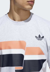 Šedé bavlněné tričko s výraznými, překrývajícími se vzory v broskvové, námořnické a bílé barvě, s černým logem Adidas na hrudi.