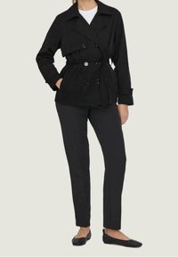 Veste noire à double boutonnage avec ceinture à nouer, grand col et manches longues, associée à un pantalon noir fuselé et des chaussures plates.