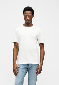 Modèle masculin aux cheveux courts portant un T-shirt blanc à manches courtes avec le logo "BOSS" et un jean bleu clair, debout devant un fond blanc uni.