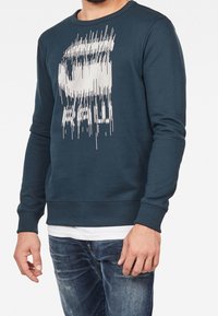 Man med mörkblå tröja med rund hals och vita, förvridna grafiska linjer och bokstäver, tillsammans med en vit undertröja och blå jeans.