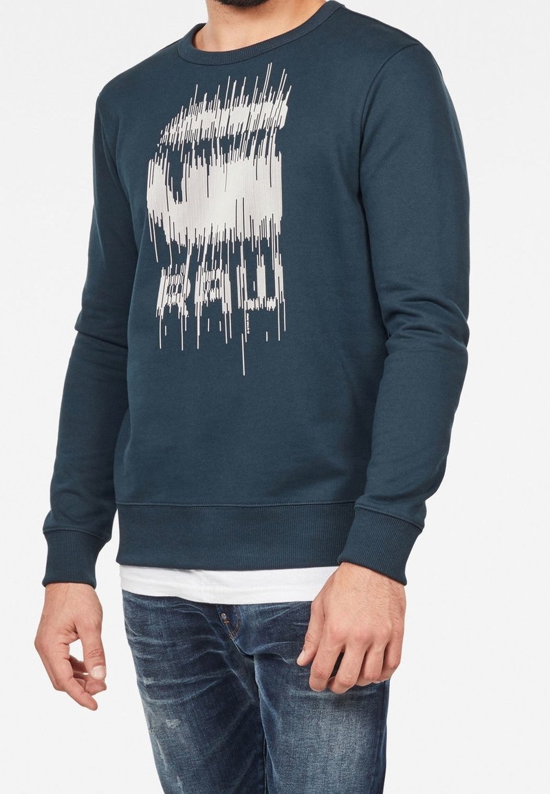 Man med mörkblå tröja med rund hals och vita, förvridna grafiska linjer och bokstäver, tillsammans med en vit undertröja och blå jeans.