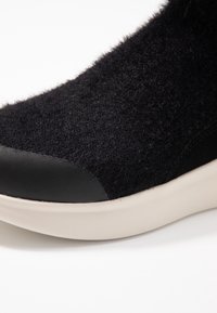 Černá teniska s měkkým, chlupatým svrškem a hladkým koženým klipem na špičce, umístěná na kontrastní bílé gumové podrážce.