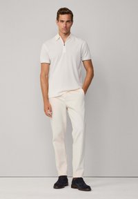Biele polo tričko so zipsom na golieri, skombinované s svetlými nohavicami. Model má na sebe tmavomodré semišové topánky. Jednoduchý, čistý dizajn.