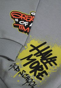 Szary sweter z odważnym graficznym napisem w czerni i pomarańczu, z żółtymi plamkami akcentującymi. Żebrowane mankiety i miękki materiał bawełniany.