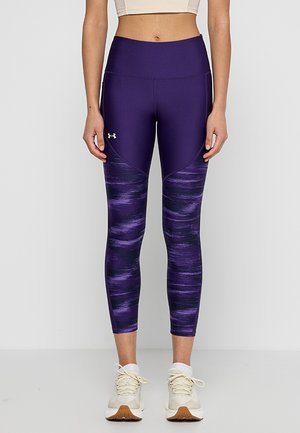 Donna che indossa leggings a vita alta di colore viola con un motivo e scarpe da ginnastica beige chiaro, in piedi contro uno sfondo bianco semplice.