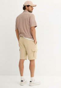 Kurzärmliges, hellbraunes Hemd mit lockerer Passform, kombiniert mit beigen Cargo-Shorts mit Seitentaschen und strukturiertem Stoff.