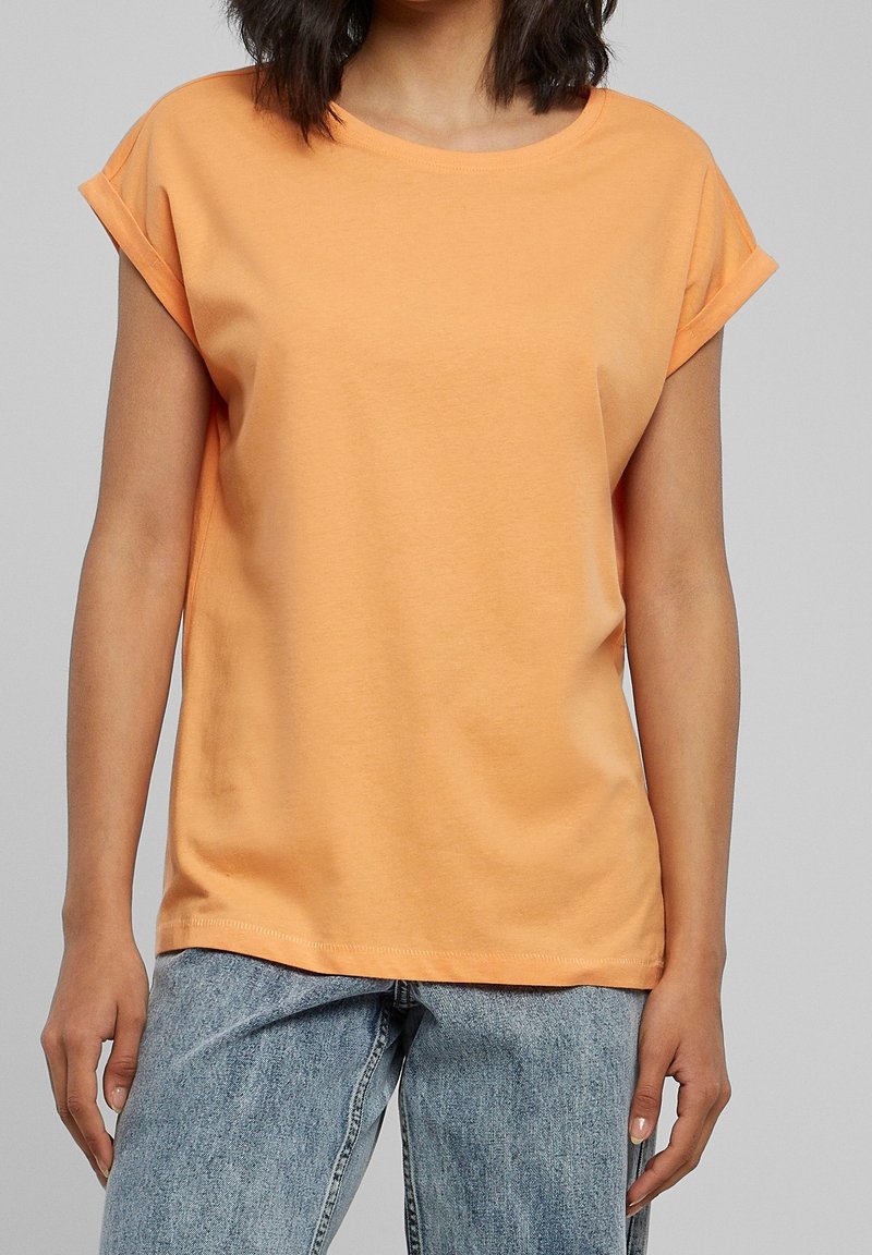 Femme portant un T-shirt uni pêche à manches courtes avec des revers retroussés et un jean en denim bleu clair, debout devant un fond neutre.