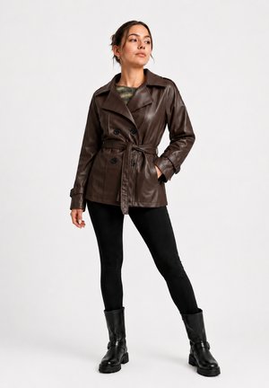 Frau trägt dunkelbraune, taillierte Lederjacke, schwarze Leggings und schwarze Stiefel und posiert vor einem schlichten hellen Hintergrund.
