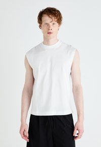Tricou alb fără mâneci din bumbac, cu guler rotund și o croială lejeră, asortați cu pantaloni scurți negri. Textură fină, design minimal.