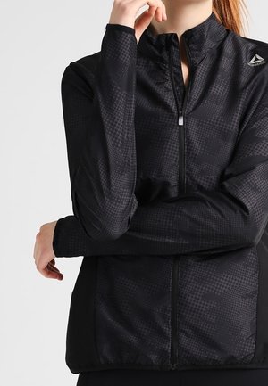 Personne portant une veste zippée noire à motifs de Reebok, un bras croisé et l’autre main près du menton, sur un fond uni.