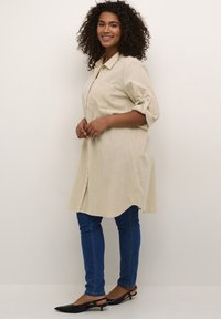 Chemise longue beige avec un col, manches retroussées et une poche poitrine, associée à un jean skinny bleu et des ballerines noires à bout pointu.