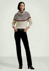 Moteris su trumpais juodais plaukais, vilkinti raštuotą grietinės spalvos megztinį su raudonais ir juodais raštais, juodas kelnes, juodą diržą ir juodas aukštakulnius batus.