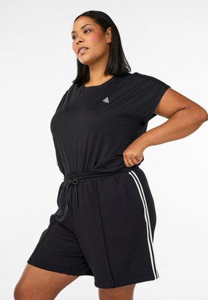 Frau, die ein schwarzes T-Shirt anpasst und dazu passende schwarze Shorts mit weißen Seitenstreifen vor einem schlichten weißen Hintergrund trägt.